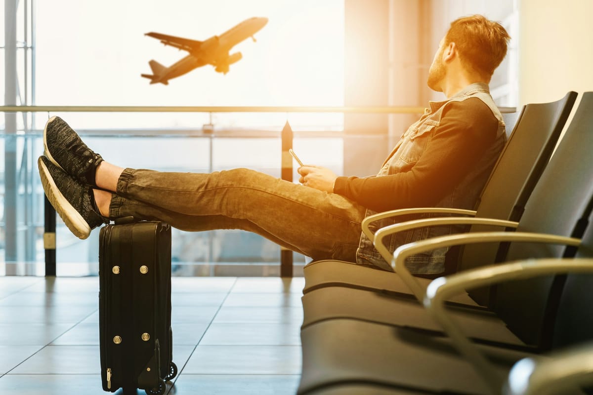 Man in a denim jacket resting his sneakers on a black carry-on suitcase in an airport gate area, watching a plane take off through the window