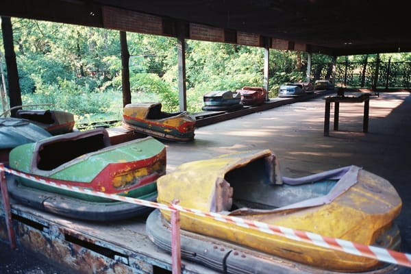 Broken, faded bumper cars lined up under a roof behind red and white warning tape, with trees in the background