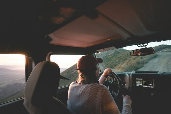 A woman in a red cap driving a 4x4 along a dirt road on a green mountainside at sunset, with the dashboard screen playing audio