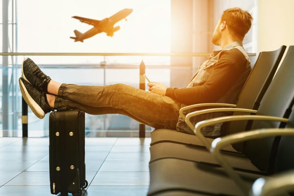 Man in a denim jacket resting his sneakers on a black carry-on suitcase in an airport gate area, watching a plane take off through the window