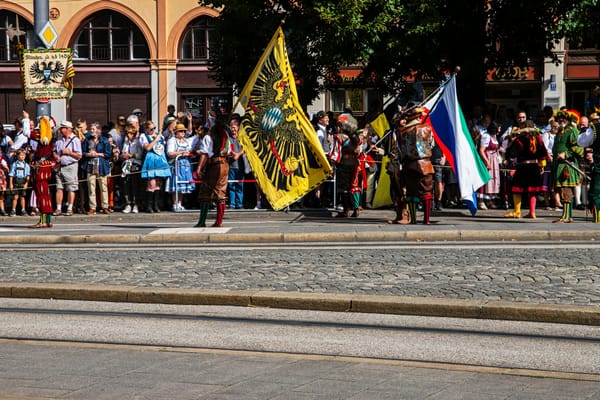 Costumed marchers with flags and traditional Bavarian dress walk past a crowd of onlookers during a festival parade on a city street