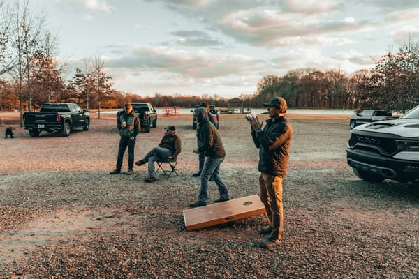 Friends playing cornhole on a gravel lot beside parked pickup trucks at sunset, one man holding a bean bag and another seated in a camp chair