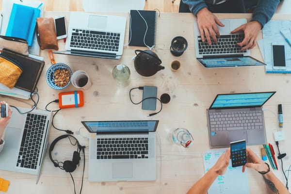 Overhead view of a shared wooden table with laptops, phones, notebooks, headphones and tea cups as people work